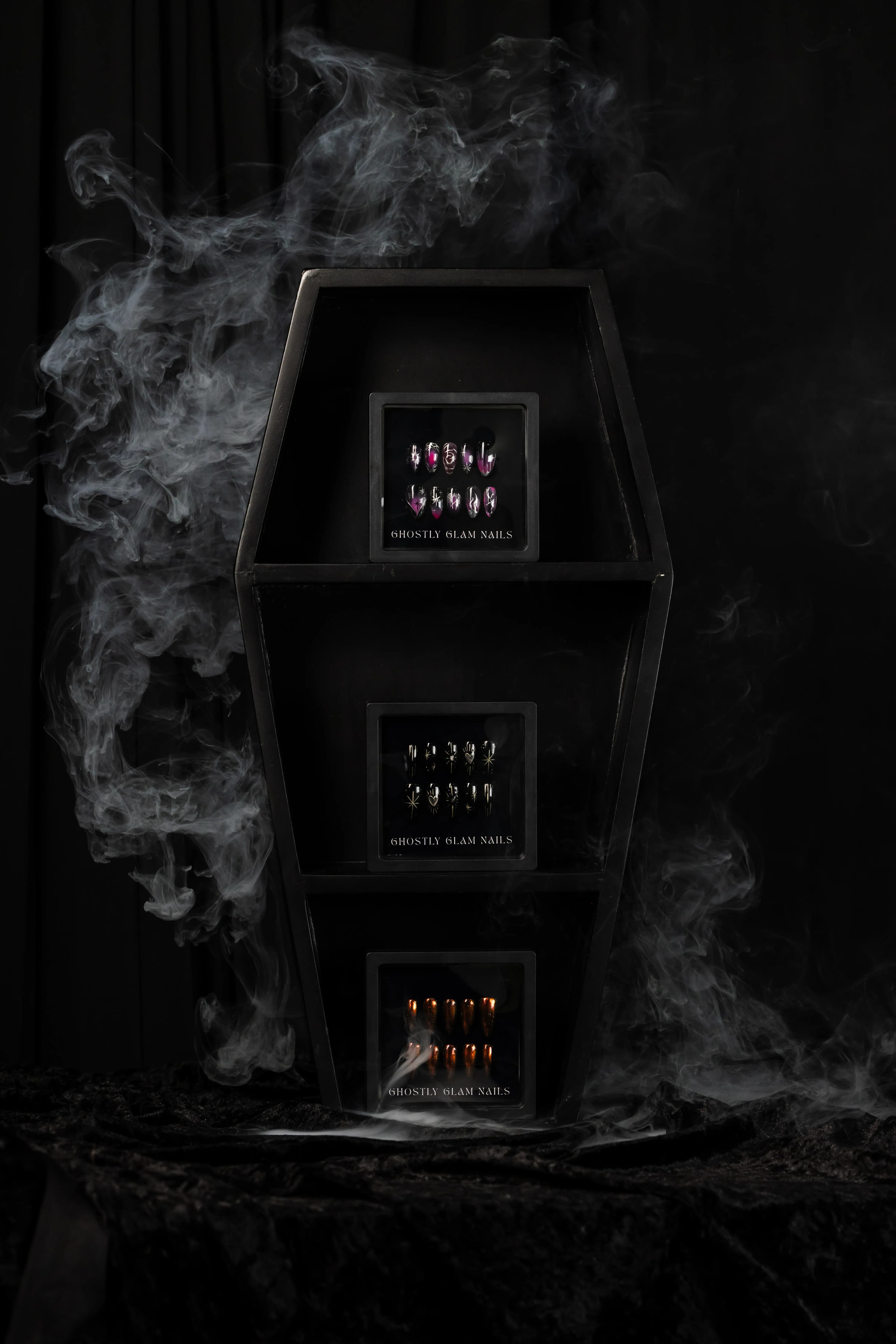 Coffin-shaped wooden shelf displaying three custom press-on nail sets, framed by drifting smoke.